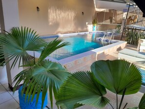 Una piscina con borde de azulejos, rodeada de macetas con plantas y un patio iluminado por el sol.