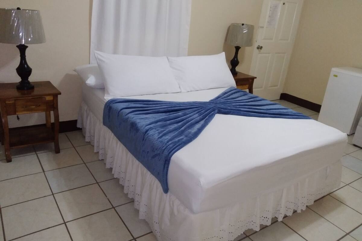 Un lit bien fait, recouvert d’une couverture bleue, deux tables de chevet avec des lampes, une porte blanche et un radiateur blanc.
