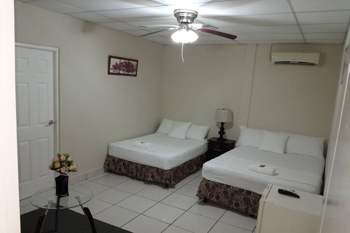Une chambre d’hôtel avec deux lits simples, une petite table avec des chaises, un ventilateur de plafond, un appareil de climatisation et une porte.