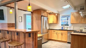A modern kitchen with wooden cabinets, a stainless steel refrigerator, and a bar area with stools. - Sunny Mid-Century Gem (Arcata)