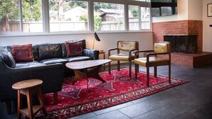 A living room with a black leather sofa, two wooden chairs, a small round table, and a red patterned rug. There is a fireplace and a TV mounted on the wall. - Sunny Mid-Century Gem (Arcata)