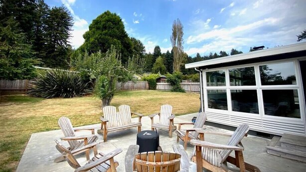 A backyard with a patio area featuring Adirondack chairs and a round table, surrounded by a wooden fence and trees. - Sunny Mid-Century Gem (Arcata)