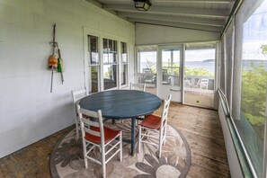En overdækket terrasse med et rundt bord og fire stole, en hvid dør, og udsigt til det fri.