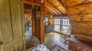 L’intérieur d’un chalet en bois, avec un lit, une commode, un fauteuil à bascule et une vue sur l’extérieur grâce à de grandes fenêtres.