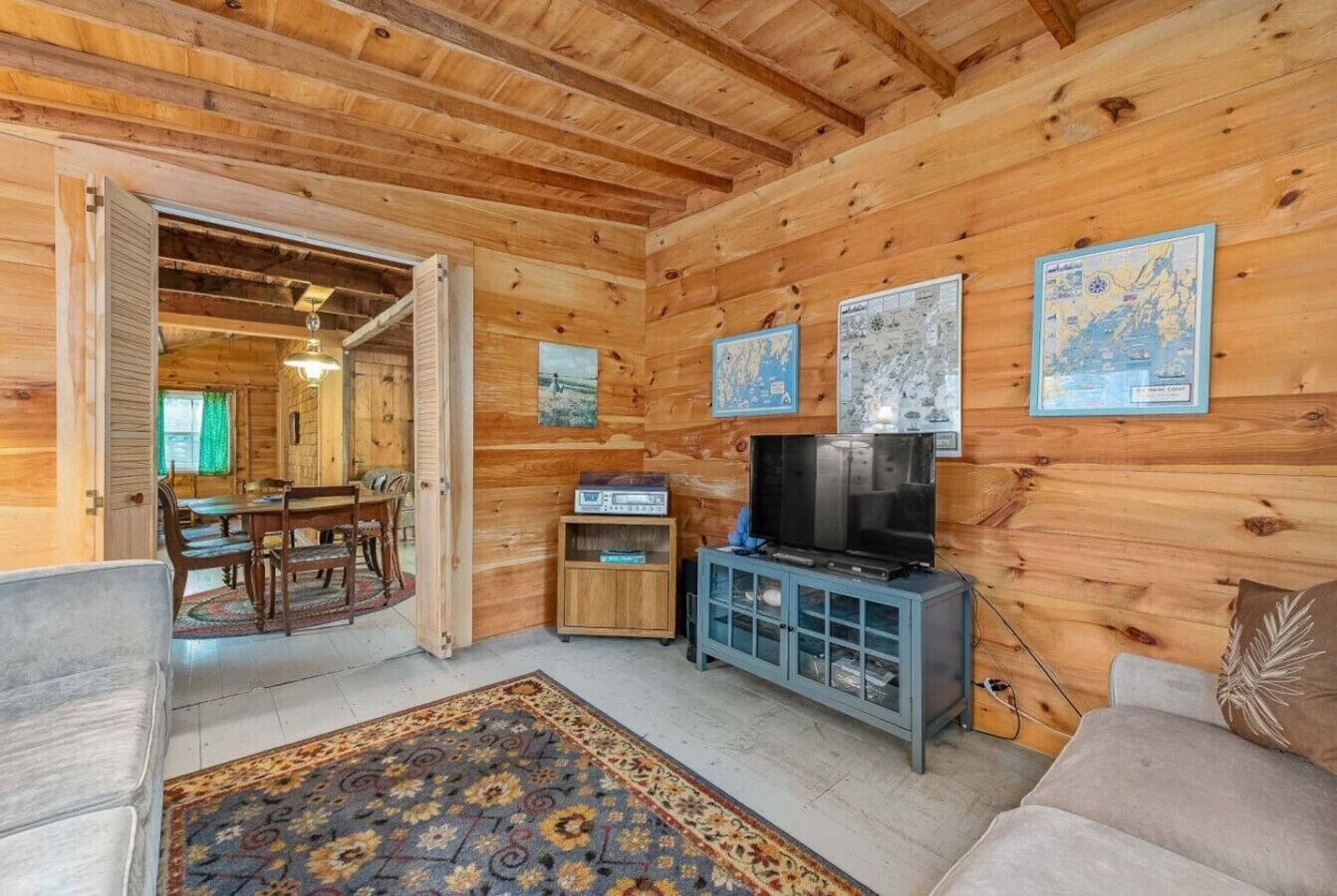 A wooden cabin interior with a dining area, a television, and a patterned rug.