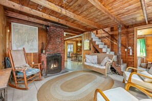A rustic interior with a brick fireplace, wooden beams, a staircase, and a dining area.