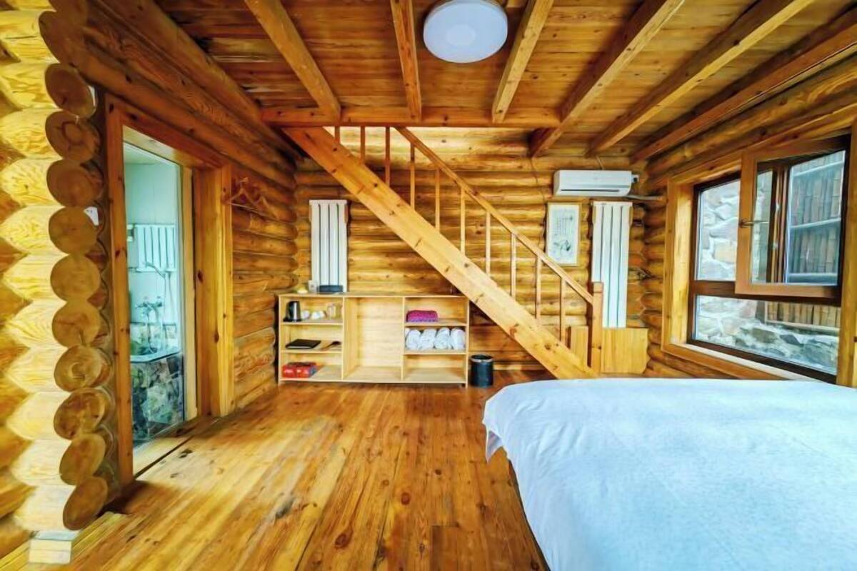 A log cabin interior with a wooden staircase, a bed, a shelf with items, and a window.