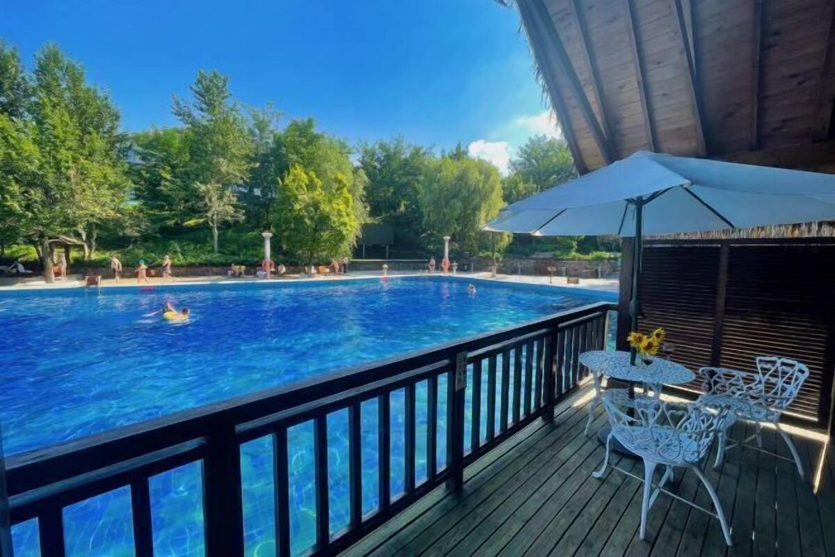 A swimming pool with a wooden deck, surrounded by trees and a covered area with white metal chairs.