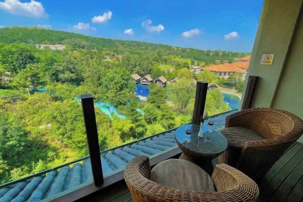 A balcony with wicker chairs and a table, overlooking a lush landscape with a swimming pool and houses.