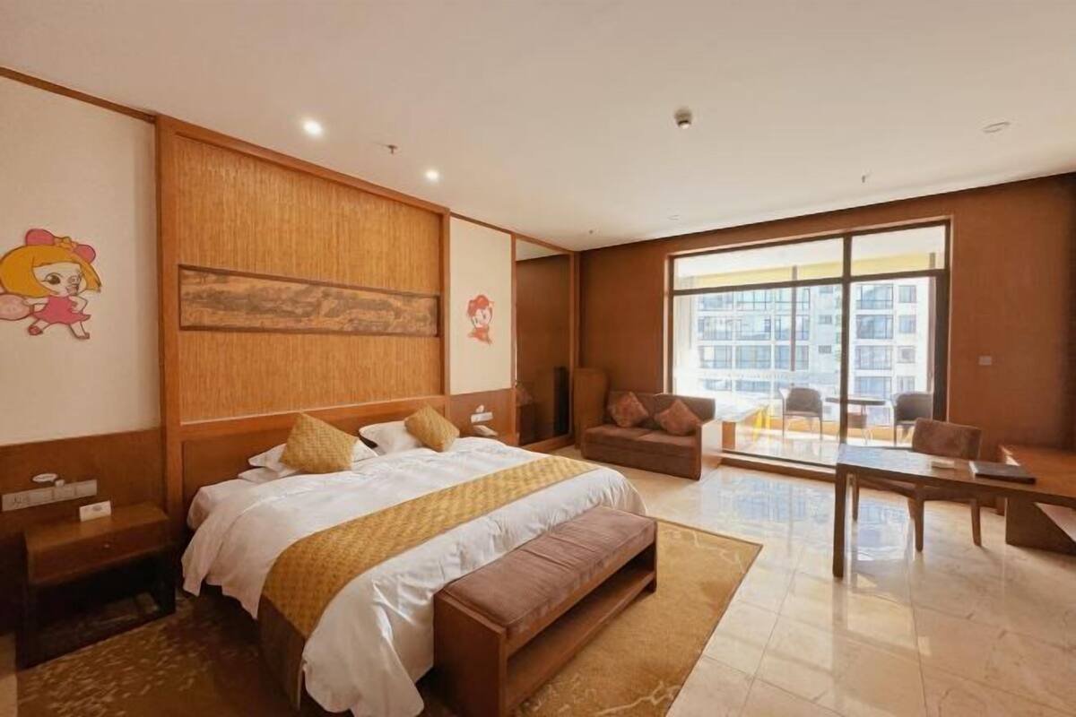 A hotel room with a large bed, a sofa, a dining table, and a view of a building through a large window.