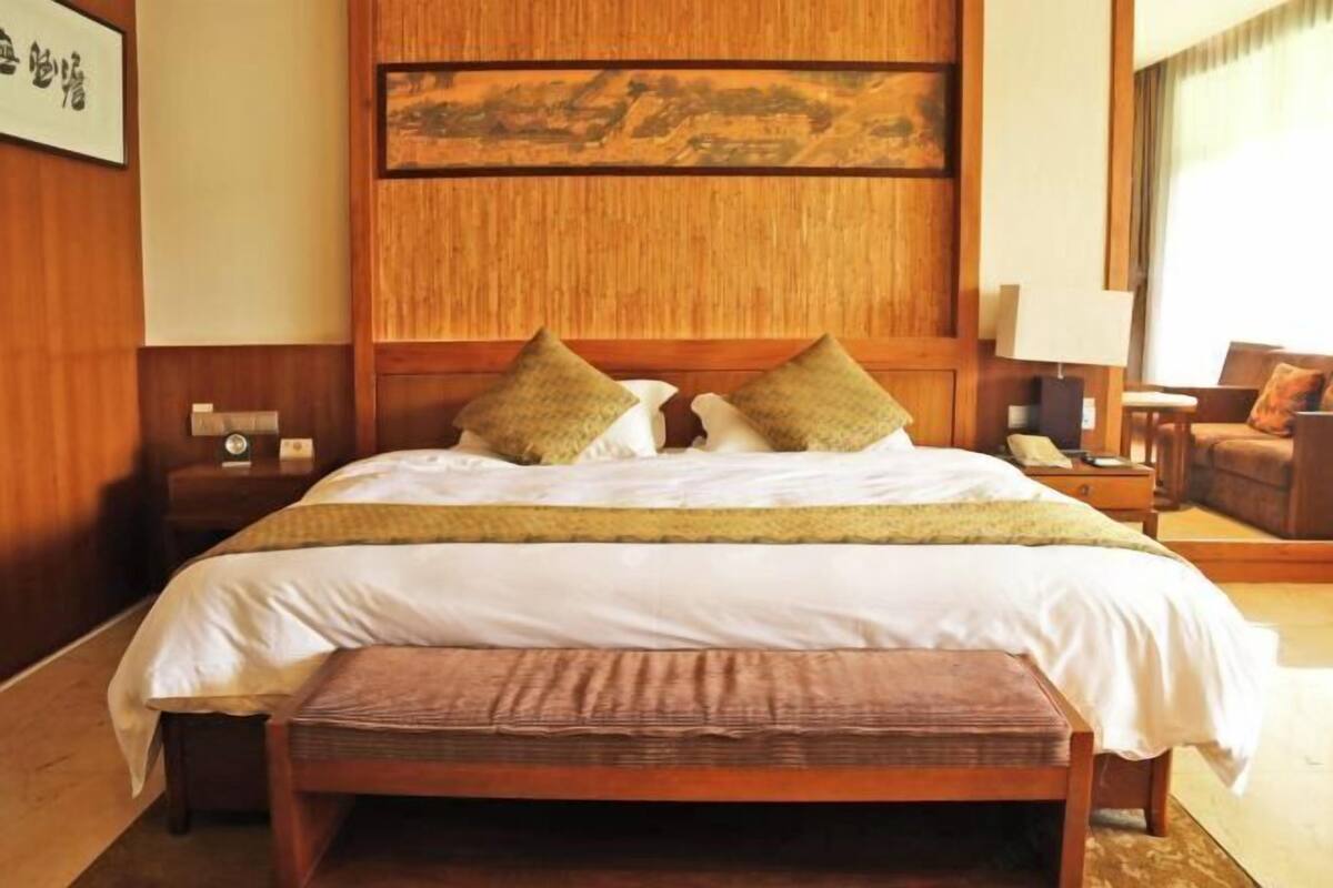 A hotel room with a wooden headboard, a bed with white bedding, two pillows, a bedside table with a lamp, and a chair.