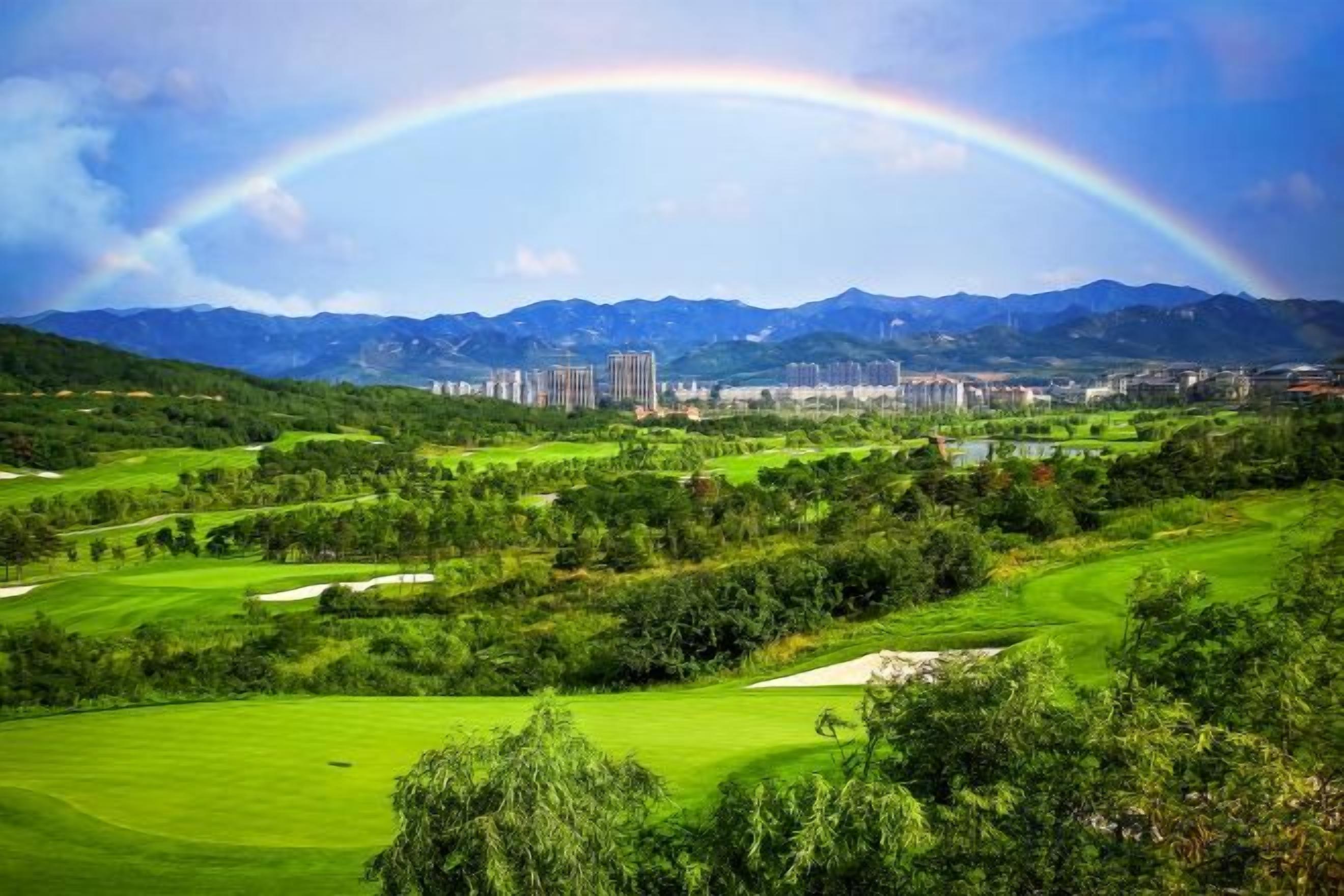 A golf course with a rainbow, mountains, and buildings in the background.