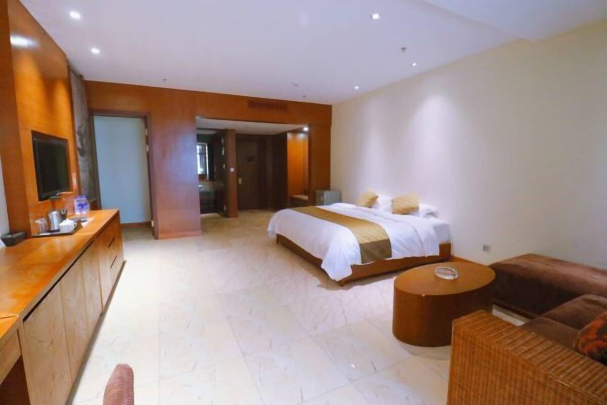 A hotel room with a bed, a sitting area with a sofa and a round table, a wooden desk with a TV, and a bathroom visible through an open door.