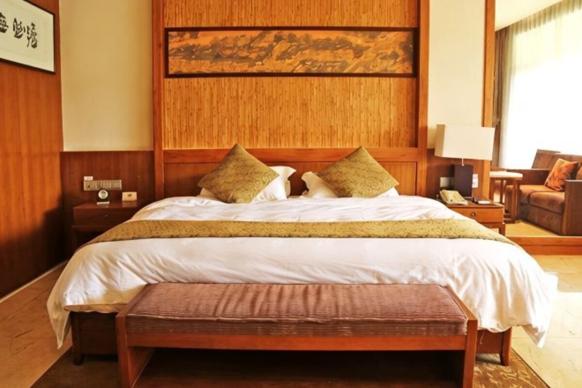 Una habitación de hotel con una cama grande, un cabecero de madera, una mesita de noche con lámpara y un pequeño rincón de descanso con una silla y una mesa.