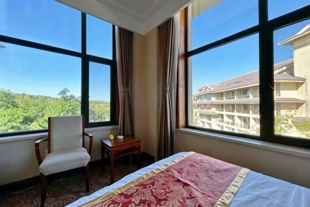 A hotel room with a bed, a chair, a small table, and large windows offering a view of a building and greenery.