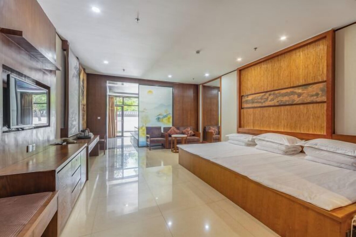 A modern hotel room with a large bed, wooden headboard, a TV, and a seating area with a view of the outdoors.
