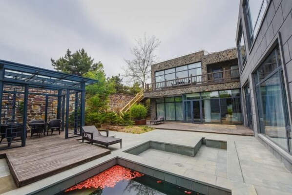 A modern outdoor patio area with a stone wall, a wooden deck, and a small pond with red flowers.