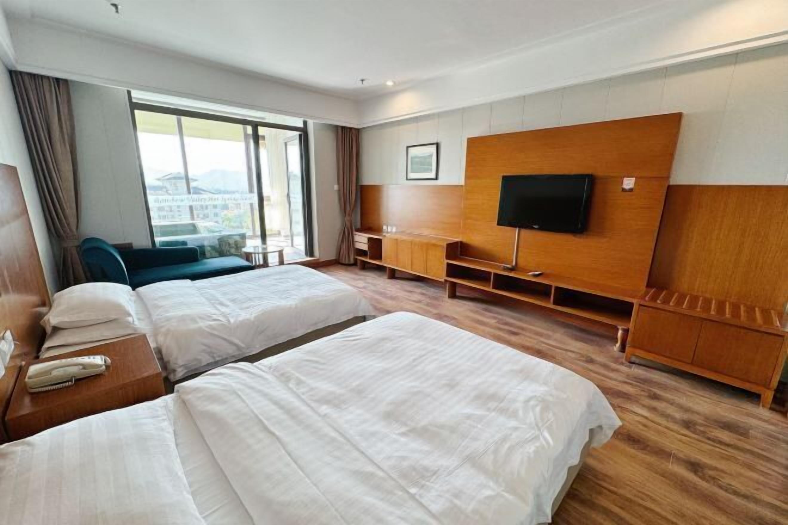 A hotel room with two beds, a TV mounted on the wall, a wooden cabinet, and a large window with curtains.