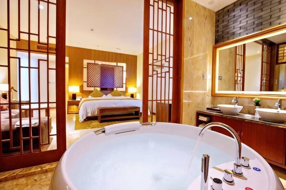 A hotel room with a large bathtub, a bed with a wooden headboard, a large mirror, and a wooden partition.