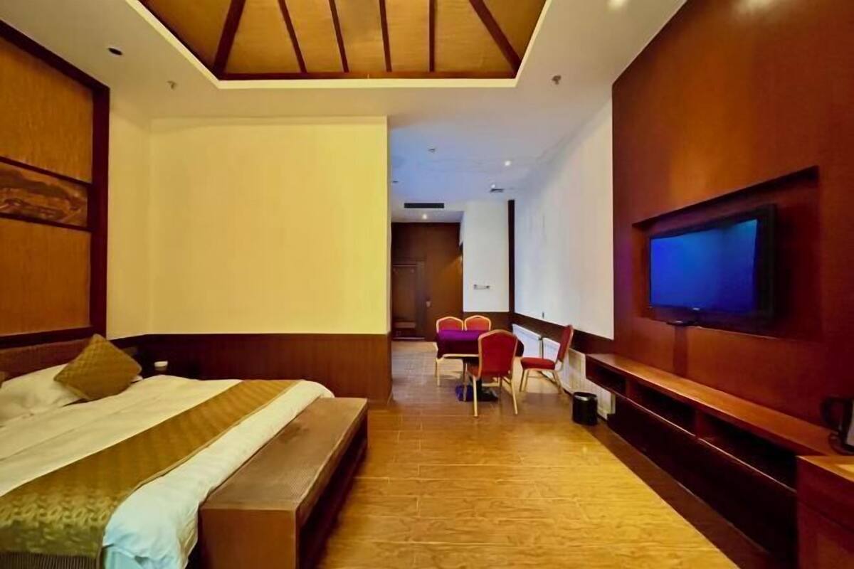 A hotel room with a bed, a TV mounted on the wall, a desk with chairs, and a wooden bench.