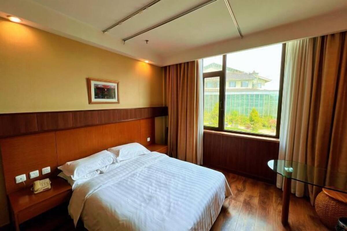 A hotel room with a bed, a nightstand, a glass table, a window with curtains, and a framed picture on the wall.