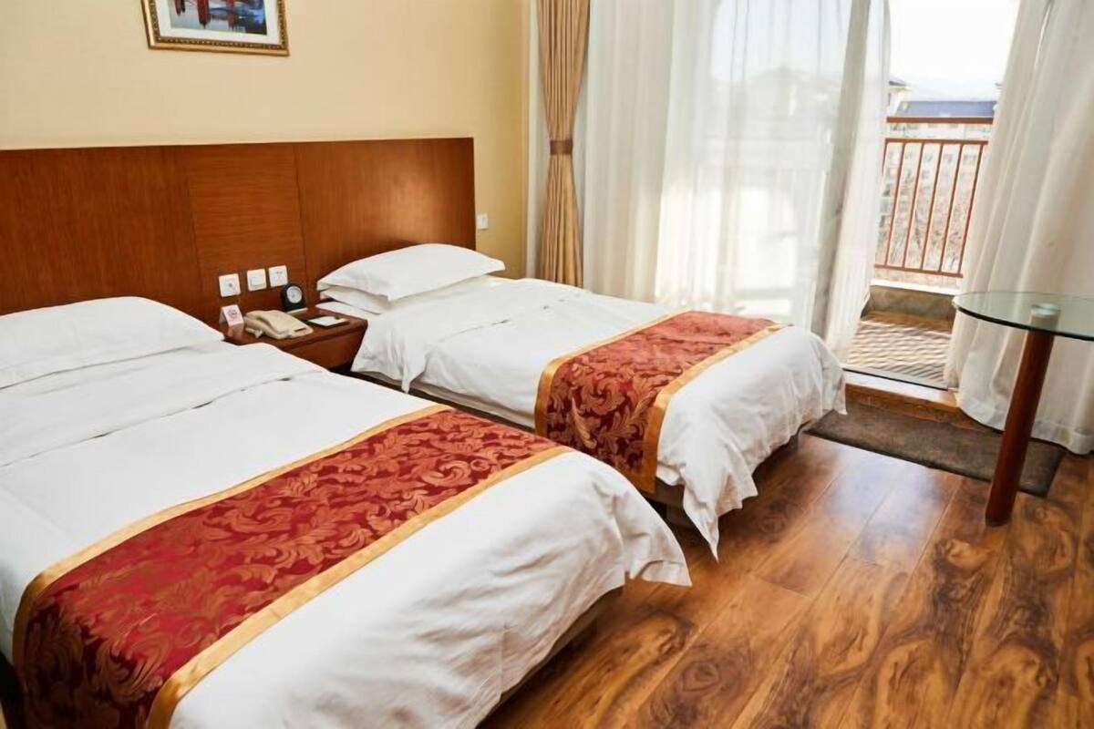 A hotel room with two beds, wooden flooring, a nightstand with a clock, and a balcony with a view.