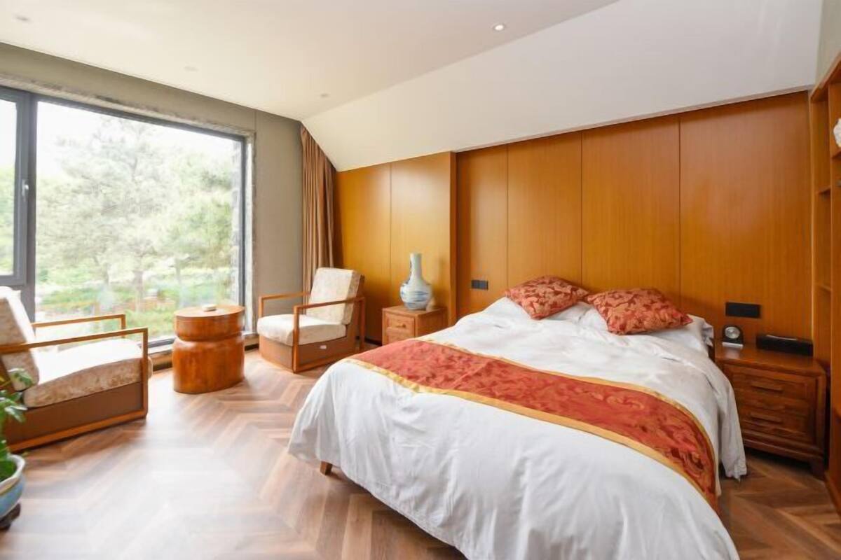 A bedroom with a large bed, a chair, a nightstand, a vase, and a window with a view of trees.