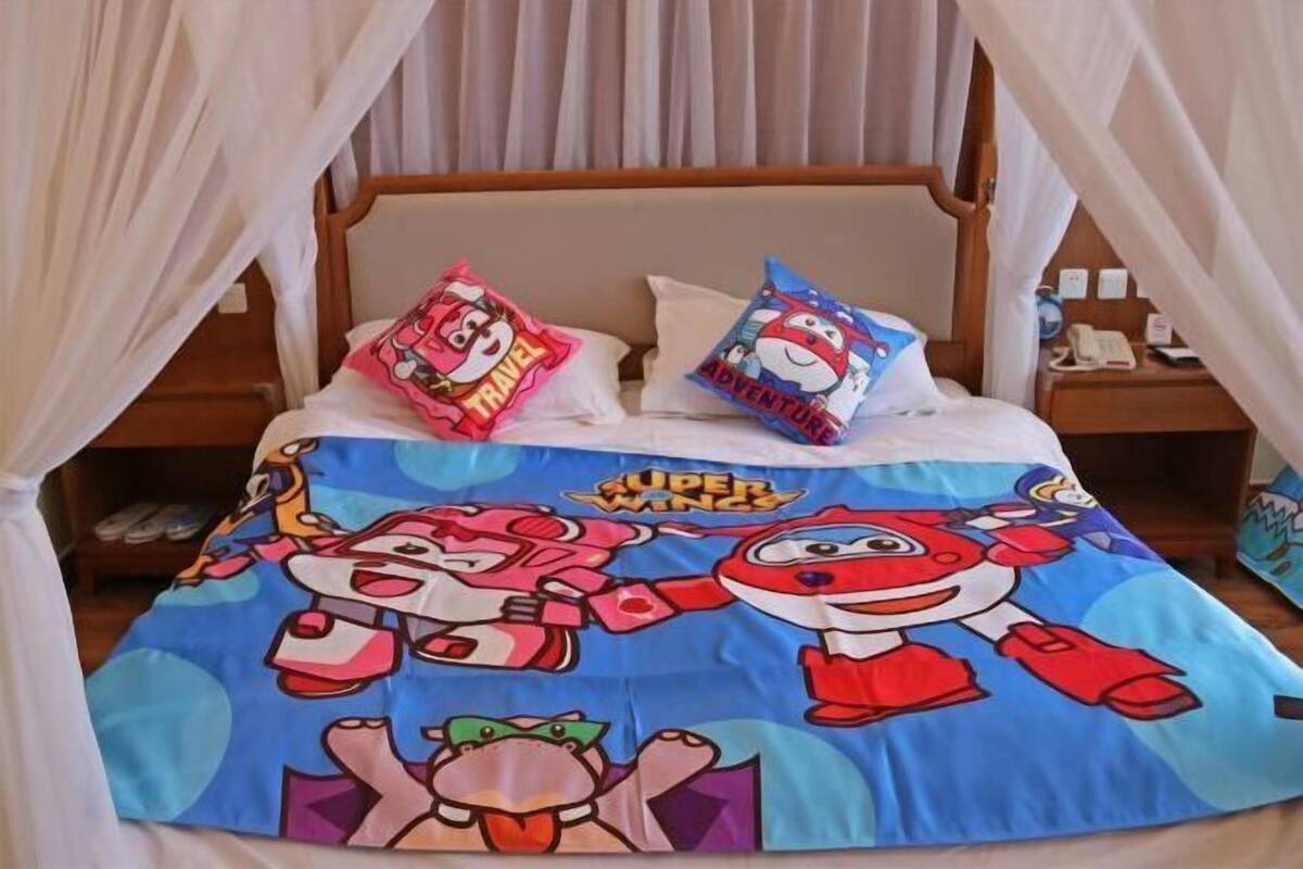 A bed with a Super Wings themed bedspread, two pillows, a wooden headboard, and a nightstand with a phone and a lamp.