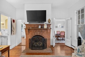 A living room with a fireplace, a television mounted above it, and a dining area visible through an open door. - Recently Renovated Home Minutes from Ocean Point (Boothbay)
