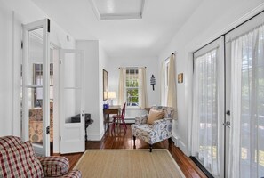 A room with a wooden floor, a patterned armchair, a dining table with chairs, and a window with curtains. - Recently Renovated Home Minutes from Ocean Point (Boothbay)