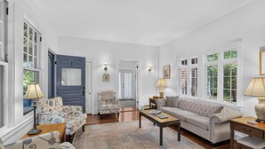 Un salon spacieux comprenant un canapé, des fauteuils, une table basse et un tapis. Deux lampes sont posées sur des tables d’appoint, et une porte donne accès à une autre pièce.