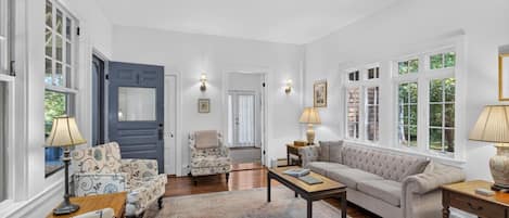 A spacious living room with a sofa, armchairs, a coffee table, and a rug. There are two lamps on side tables, and a door leading to another room.