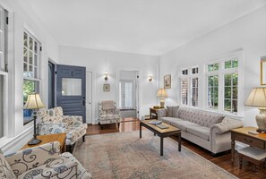 Ein geräumiges Wohnzimmer mit einer Couch, Sesseln, einem Couchtisch und einem Teppich. Auf Beistelltischen stehen zwei Lampen, und eine Tür führt in einen anderen Raum.