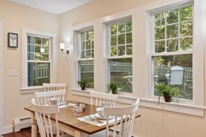 A dining room with a wooden table and white chairs, large windows with white frames, and potted plants on the windowsills. - Recently Renovated Home Minutes from Ocean Point (Boothbay)