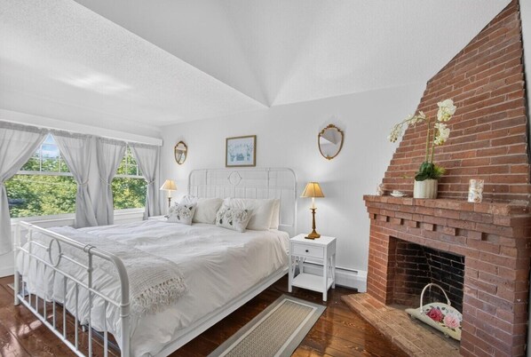 A bedroom with a white metal bed, a white nightstand, a brick fireplace, and a window with curtains. - Recently Renovated Home Minutes from Ocean Point (Boothbay)