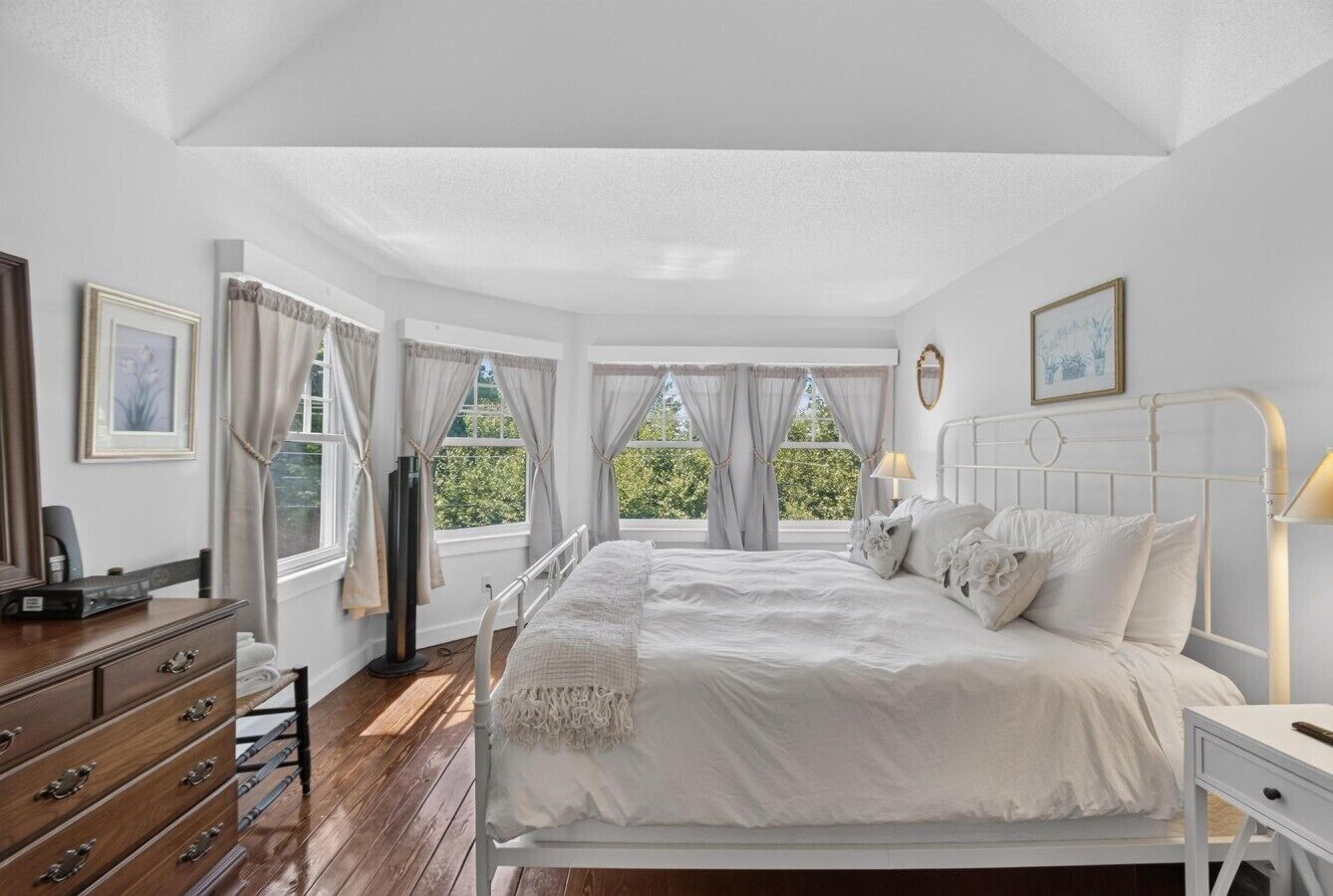 Un dormitorio con cama, mesitas de noche, cómoda y ventanas con cortinas.