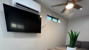 A modern living room with a flat-screen TV mounted on the wall, a ceiling fan, and a wooden dining table with benches. - West University Carriage Home & Parking (Tucson)