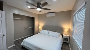 A bedroom with a bed, two bedside tables, a ceiling fan, a window with blinds, and a door. - West University Carriage Home & Parking (Tucson)