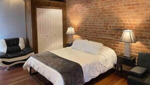 Een slaapkamer met een bakstenen muur, een bed met wit beddengoed, twee bedlampen, een stoel en een garderobe.