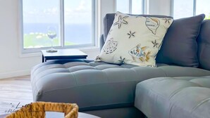 A modern living room with a grey sectional sofa, a decorative pillow with sea-themed designs, and a view of the ocean through large windows.