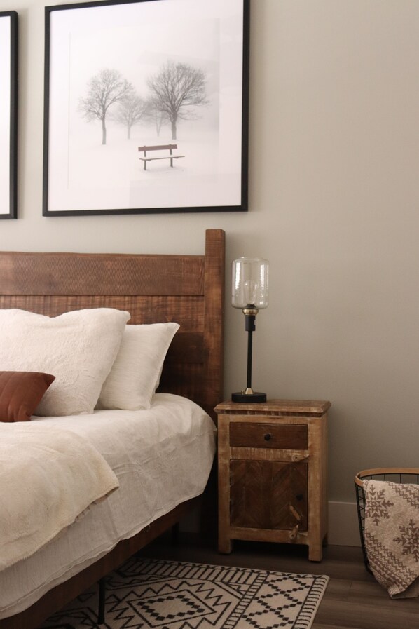 A neatly made bed with white bedding and a brown pillow, a wooden bedside table with a lamp, a framed picture on the wall, and a patterned rug on the floor.