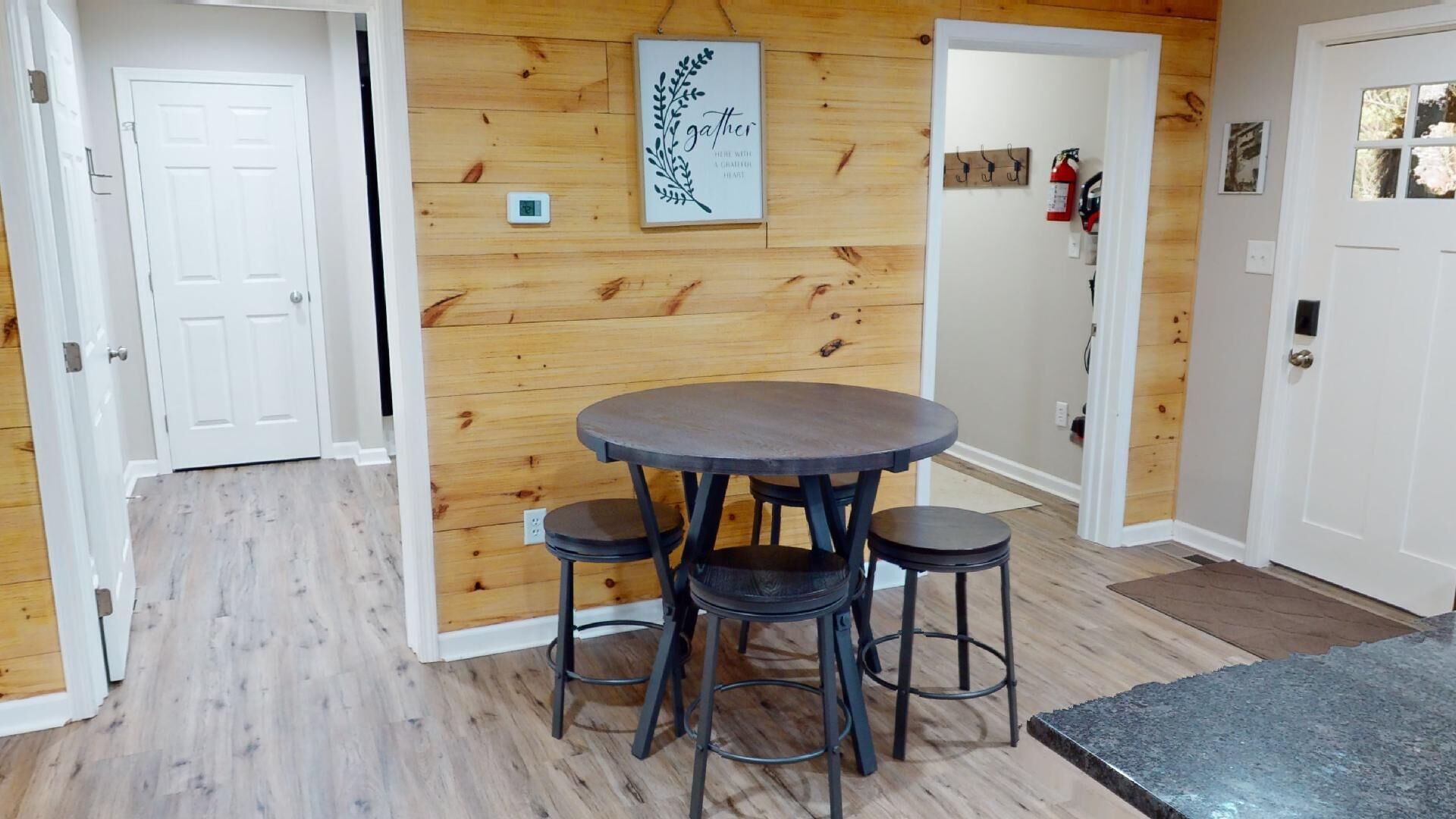 A room with a round table and four stools, wooden walls, a white door, and a small framed artwork.