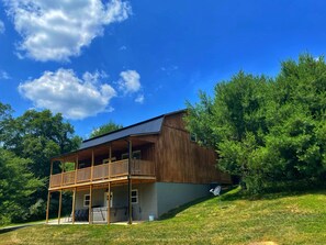 A two-story wooden house with a balcony, surrounded by trees and a clear sky.