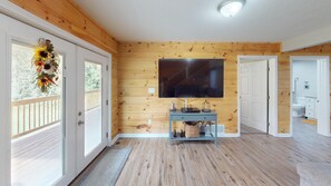 A living room with a wooden paneled wall, a flat-screen TV mounted above a blue console table, and a view of the adjacent bathroom through an open door.
