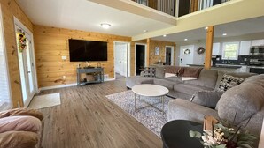 A spacious living room with a large sectional sofa, a flat-screen TV mounted on the wall, and a wooden floor.