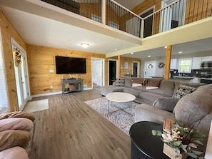 A spacious living room with a large sectional sofa, a flat-screen TV mounted on the wall, and a wooden floor.