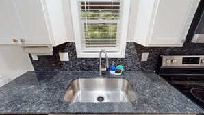 A kitchen with a stainless steel sink, a window with blinds, and a black tiled backsplash.