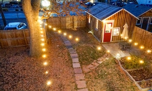 Une petite cabane en bois avec une porte rouge, entourée d’un chemin de pierres et illuminée par des guirlandes lumineuses.