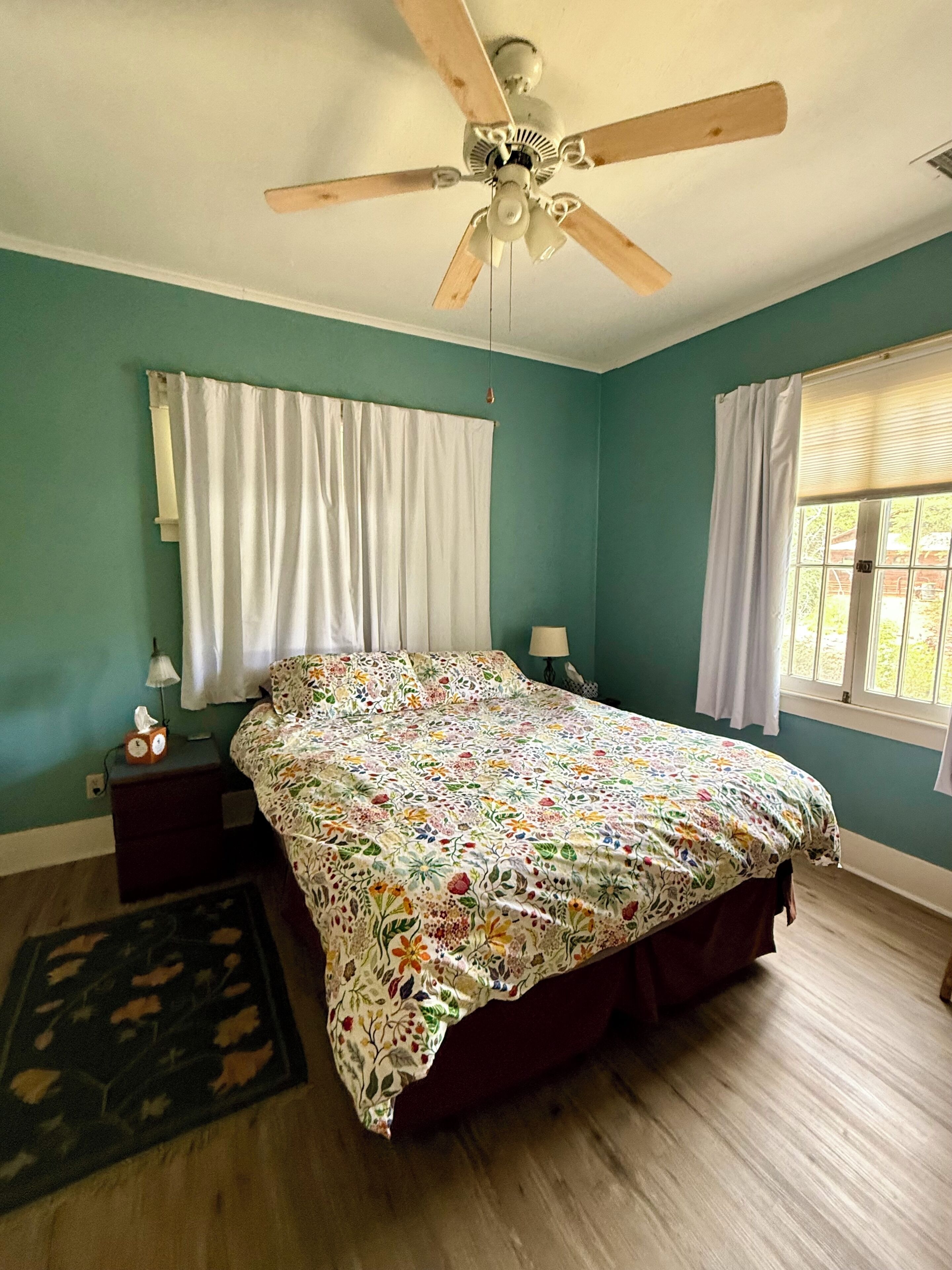 Ein Schlafzimmer mit Deckenventilator, einem Bett mit floralem Überwurf, zwei Nachttischlampen, einem Fenster mit Vorhängen und einem Nachttisch mit einer Uhr.