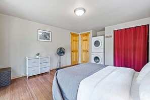 Een slaapkamer met een bed, een wasmachine, een ventilator en een dressoir.